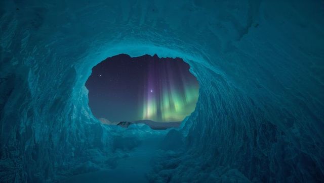 Framing oval ice cave opening revealing northern lights over snow-covered hills