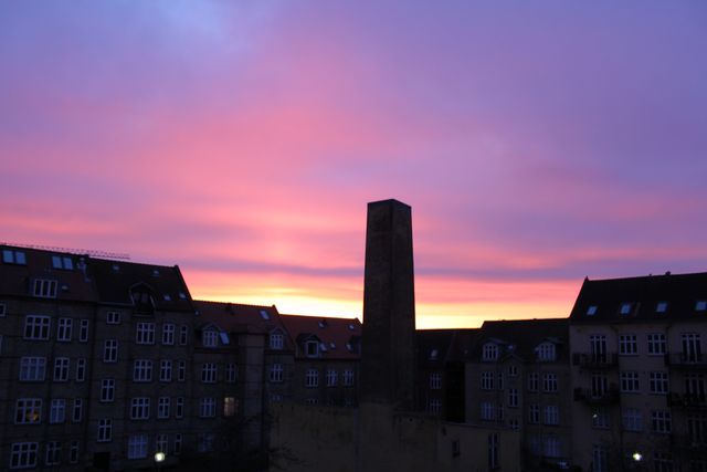 This image shows a colorful sunset sky with a striking mix of purple and pink hues over urban buildings. A tall tower stands silhouetted against the vibrant backdrop. This can be used for urban photography themes, artistic backgrounds, or promotional materials for travel and city-based content.