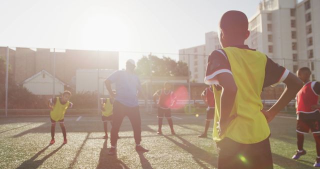 Young football players warming up under coach's guidance at an urban sports facility with sunshine lighting up the scene. Ideal for themes of team building, fitness, outdoor activities, youth sports programs, healthy lifestyle initiatives.
