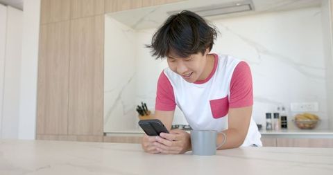 Asian Student Smiling at Smartphone in Modern Kitchen