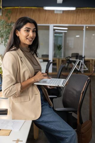 Indian professional woman holding laptop and reviewing charts while leaning on desk