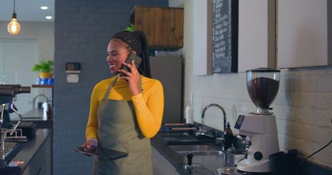 African american barista talking on phone holding tablet at modern cafe counter with grinder