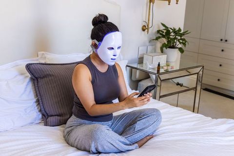 Woman wearing led facial mask relaxing on bed using smartphone for at-home skincare