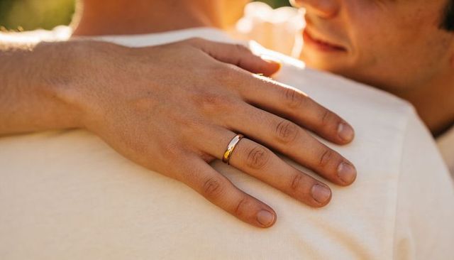 Closeup hand resting on partner's shoulder wearing gold wedding band during golden-hour embrace