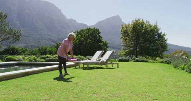 An older woman is seen gardening on a sunny day in a scenic yard with a backdrop of mountains and lush greenery. This image is perfect for articles or advertisements related to gardening, active lifestyles for seniors, outdoor relaxation, or retirement living. It conveys a sense of peaceful enjoyment in nature, ideal for promoting healthy living and leisure activities.