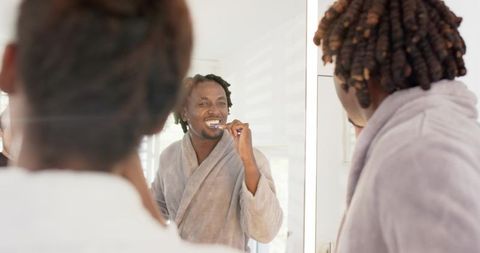 African couple brushing teeth together at mirror, man in gray robe smiling morning routine