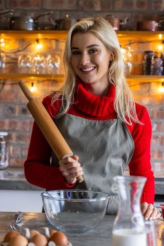 Blonde woman baking in cozy rustic kitchen holding rolling pin and smiling by warm lights