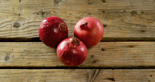 Three red onions rest on a rustic wooden surface, showcasing their vibrant color and texture. Their placement against the weathered wood highlights the natural simplicity of fresh produce.