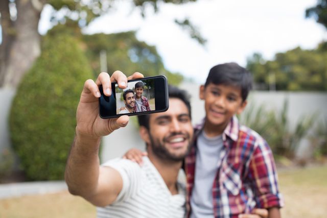 Father and son smiling while taking a selfie in a yard. Ideal for use in family-oriented advertisements, parenting blogs, social media campaigns, and lifestyle articles focusing on family bonding and outdoor activities.