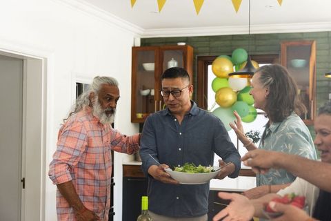 Friends hosting casual kitchen celebration sharing salad and wine among mature adults