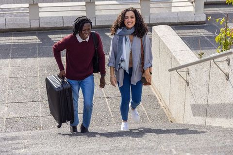 Young diverse travelers walking up urban steps pulling wheeled suitcase and smiling