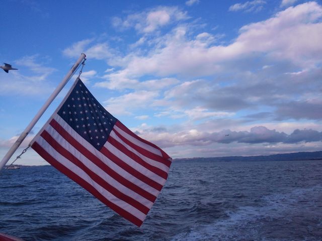 American flag prominently displayed on a boat, waving gently in the wind against a serene ocean and expansive sky with clouds. Ideal for use in contexts celebrating patriotism, travel, tourism, and natural beauty in the USA, as well as promotions related to marine activities or the Fourth of July.