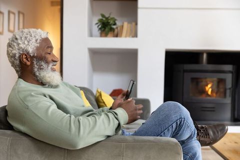 Senior african american man relaxing on sofa using smartphone by cozy fireplace smiling