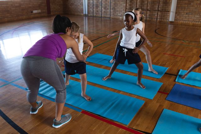 Female yoga instructor guiding children in a school gym. Children are practicing yoga on mats, learning stretching and fitness techniques. Ideal for use in educational materials, fitness programs, and wellness promotions.