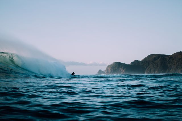 Surfer riding a large ocean wave near a breathtaking, rugged coastline. Perfect for content related to ocean adventures, extreme sports, and coastal exploration. Can be used in sports advertisements, travel brochures, and websites promoting water sports and outdoor activities.