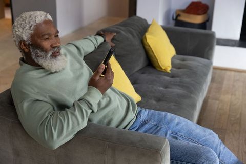 Senior african american man reclining on gray sofa using smartphone with yellow pillows