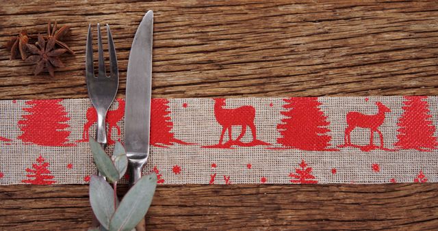 A festive table setting features a knife and fork resting on a Christmas-themed napkin with rustic charm, with copy space. The arrangement, complete with a star anise and greenery, evokes a cozy holiday atmosphere.