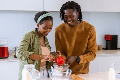 African american couple baking together in bright modern kitchen, sifting flour, smiling