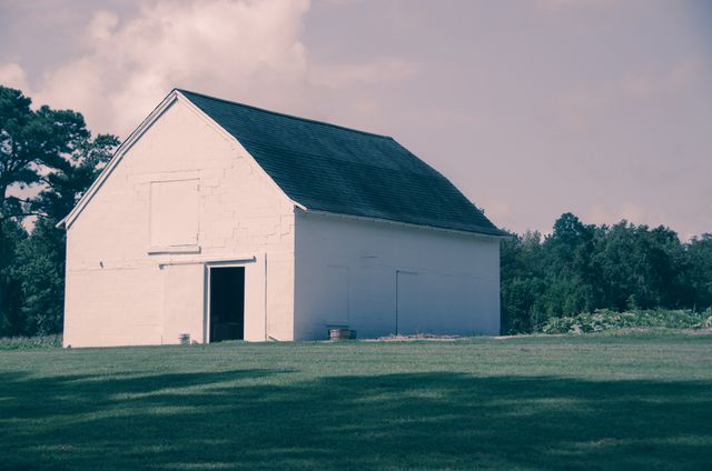 White barn stands isolated in the countryside on a sunny day, surrounded by green fields and a clear, blue sky. Ideal for using in agricultural, rural living, or real estate content. Perfect for promoting serene environments and rural retreats.