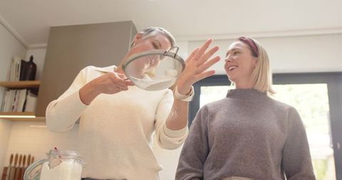 Two women sifting flour together in bright kitchen, smiling and baking together