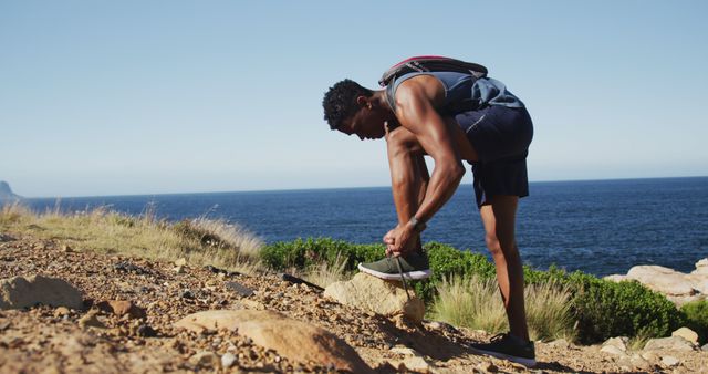 Young African American man tying shoelaces on rugged coastal trail overlooking ocean. The clear blue sky and surrounding greenery highlight the beauty of nature and the adventurous spirit of hiking. This photo can be used to promote outdoor activities, fitness, healthy lifestyles, travel, and adventure. Ideal for blogs, advertisements, and social media posts focused on active living and outdoor exploration.