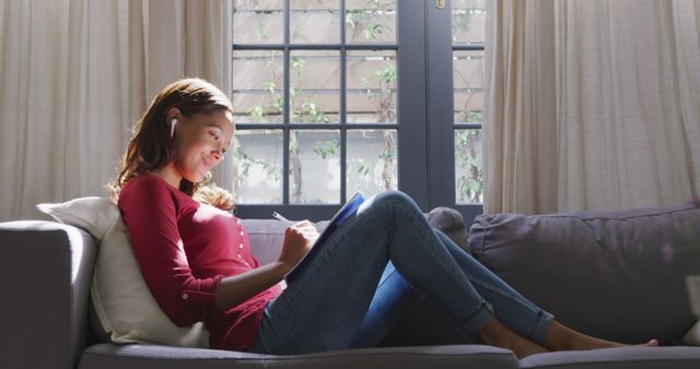 Young woman sits on sofa with earphones, listening to music while journaling in a bright living room. Perfect use for themes related to relaxation, self-reflection, home comfort, and leisure activities.
