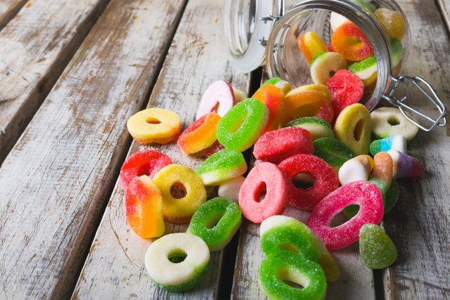 Vibrant sugar candies spilling from a glass jar onto a rustic wooden table. Perfect for illustrating concepts of indulgence, sweet treats, and unhealthy eating habits. Ideal for use in food blogs, advertisements for confectionery, or articles discussing diet and nutrition.