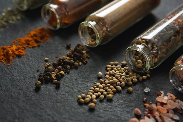 This image shows a close-up view of various spices such as coriander, pepper, salt, and turmeric spilled from glass jars onto a dark surface. Ideal for use in culinary blogs, cooking websites, recipe books, and food-related advertisements. It highlights the natural and organic aspects of cooking ingredients, making it perfect for promoting gourmet and healthy eating.