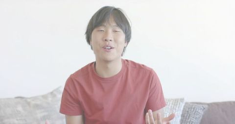 Asian man speaking and gesturing on sofa in bright minimalist living room wearing red tee