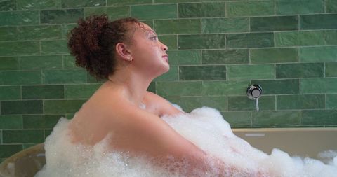 Contemplative african american woman soaking in bubble bath against green tile wall