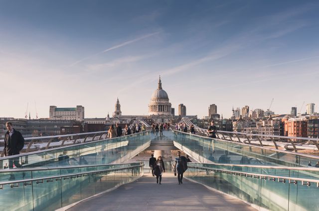 This stock photo shows people walking on Millennium Bridge with St. Paul's Cathedral in the background, under a clear sky. Perfect for use in travel blogs, articles about London, promotional material for tours, or any project celebrating famous British landmarks and cityscapes.