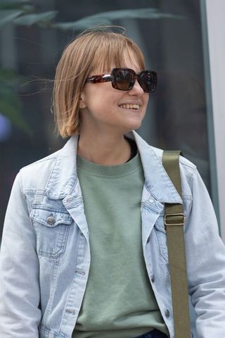 Smiling woman in denim jacket wearing tortoiseshell sunglasses carrying olive shoulder bag