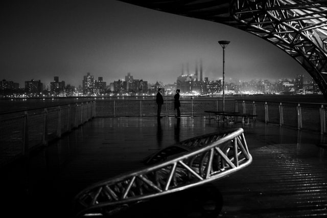 This black-and-white photo showcases two silhouetted figures standing on a waterfront deck at night, with a city's skyline illuminated in the background. The mood is serene and contemplative, perfect for urban-themed designs or conveying solitude in a bustling city. Potential uses include backgrounds for websites, blogs about urban life, or artworks evoking nighttime urban settings.