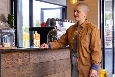Person with shaved head ordering coffee at sunlit cafe counter with pastry display
