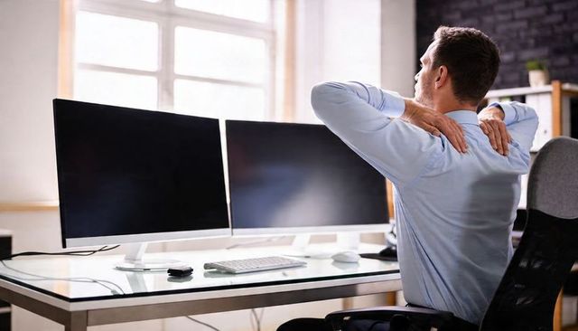 Office worker stretching and rubbing neck at glass desk with dual monitors and ergonomic chair