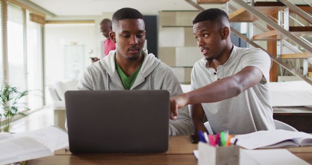 Two african american teenage twin brothers using a laptop and talking with father in background. family leisure time at home together during quarantine lockdown.