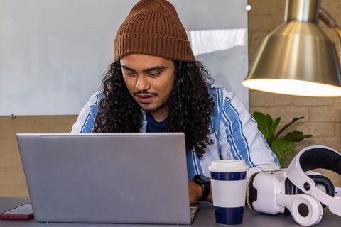 Young man wearing beanie working on laptop at home desk with vr headset and coffee
