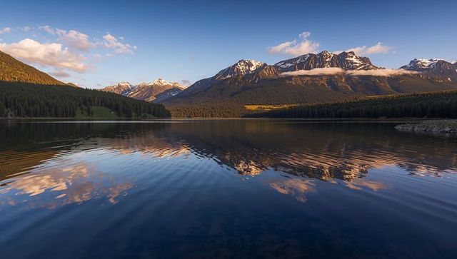 Sunrise reflection of snow-capped mountain range across alpine lake, forest shoreline