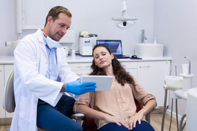 Dentist explaining treatment plan to female patient using a digital tablet in a modern dental clinic. Useful for illustrating dental consultations, patient care, modern dental technology, and professional healthcare services.