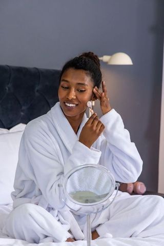 African woman in white robe using facial roller on bed for skincare routine and relaxation