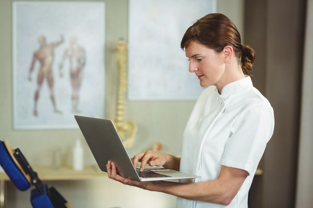 Physiotherapist in white uniform using laptop in a clinic. Background includes anatomical charts and spine model. Ideal for use in healthcare, medical technology, physiotherapy, and professional settings.