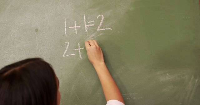 Child writing simple math equations using chalk on a classroom chalkboard. Useful for educational content, teaching resources, math tutorials, learning materials, and school-themed publications.