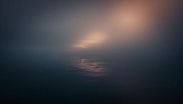 Misty dawn over tranquil sea with central shimmering light band and soft glassy ripples