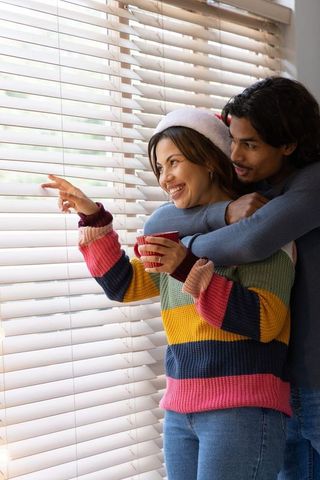 Couple hugging at window wearing santa hat and colorful knit sweater holding hot mug