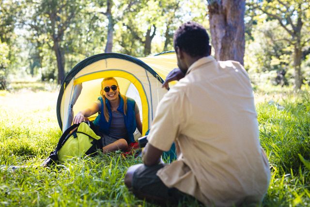 This image shows a happy, diverse couple enjoying a camping trip in a park on a sunny day. They are sitting on the grass near a tent, smiling and engaging in conversation. This photo can be used for promoting outdoor activities, travel, adventure, and lifestyle content. It is ideal for websites, blogs, and advertisements related to camping, nature, and spending quality time with loved ones.