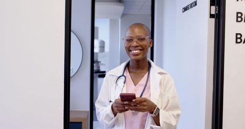 Smiling african woman doctor holding smartphone in clinic doorway wearing lab coat and stethoscope