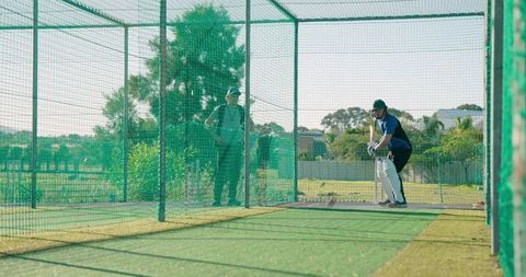 Adult cricketer practicing batting technique in outdoor net cage with coach observing