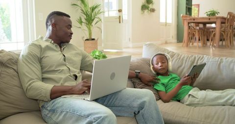 African american father working on laptop while son using tablet with headphones on sofa