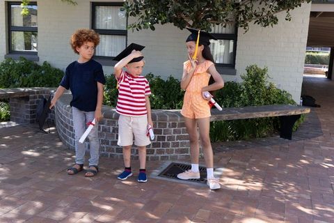 Young graduates wearing caps and holding diplomas in sunny school courtyard