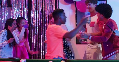 Diverse young friends celebrating around pool table under neon lights at birthday party